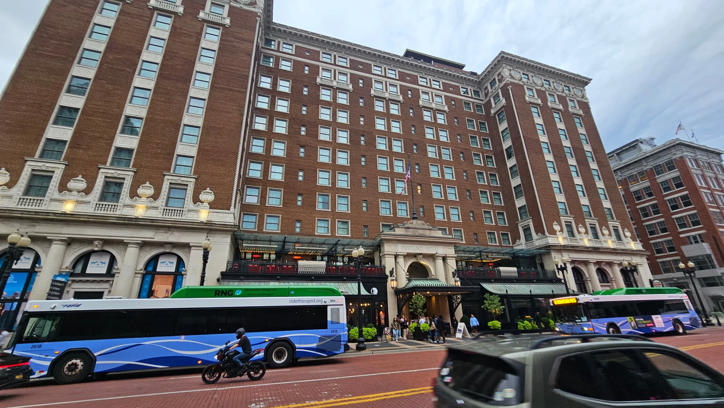 Two buses pass the entrance of a tall red-brick hotel in downtown Grand Rapids.
